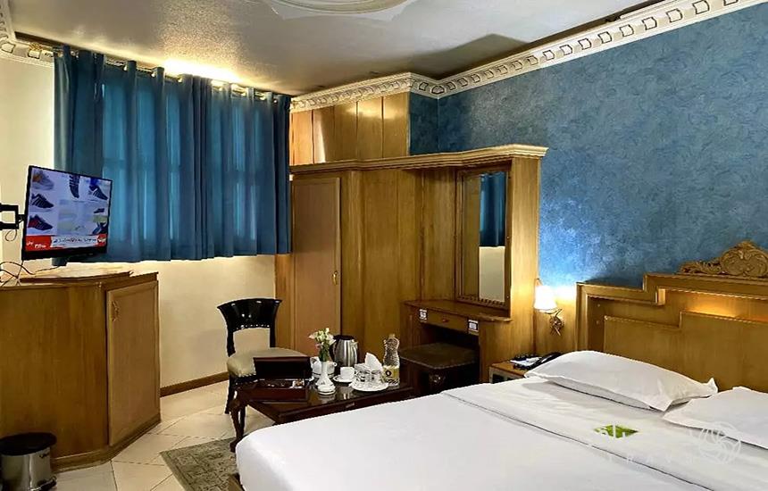 اتاقی با رنگ‌های غالب قهوه‌ای و آبی در هتل «زهره» اصفهان که تختی دونفره، یک میز و صندلی و یک مانیتور در آن است.